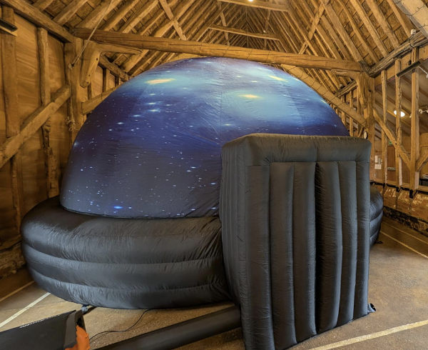 A large, dark inflatable dome with a doorway at the front. It has an image of a starfield on the dome part. It is inside a barn-like building.